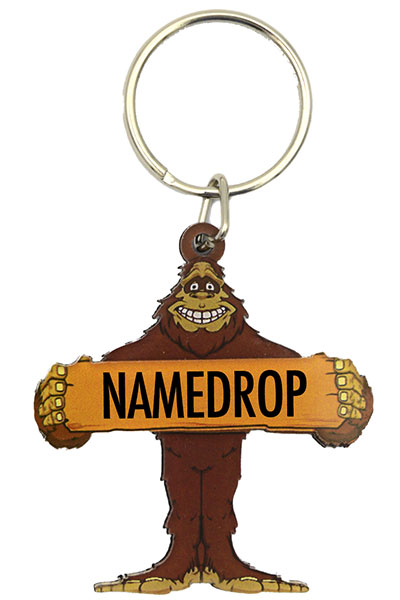 A custom wholesale keychain shaped like a smiling cartoon Bigfoot holding a wooden sign that reads NAMEDROP