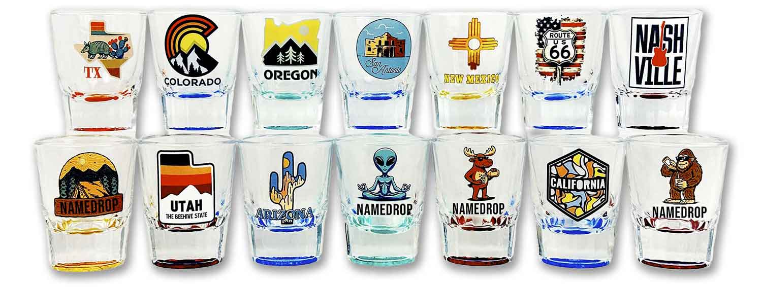 A wholesale display of clear glass shot glasses featuring colorful custom printed designs for US states and regional attractions alongside customizable namedrop samples